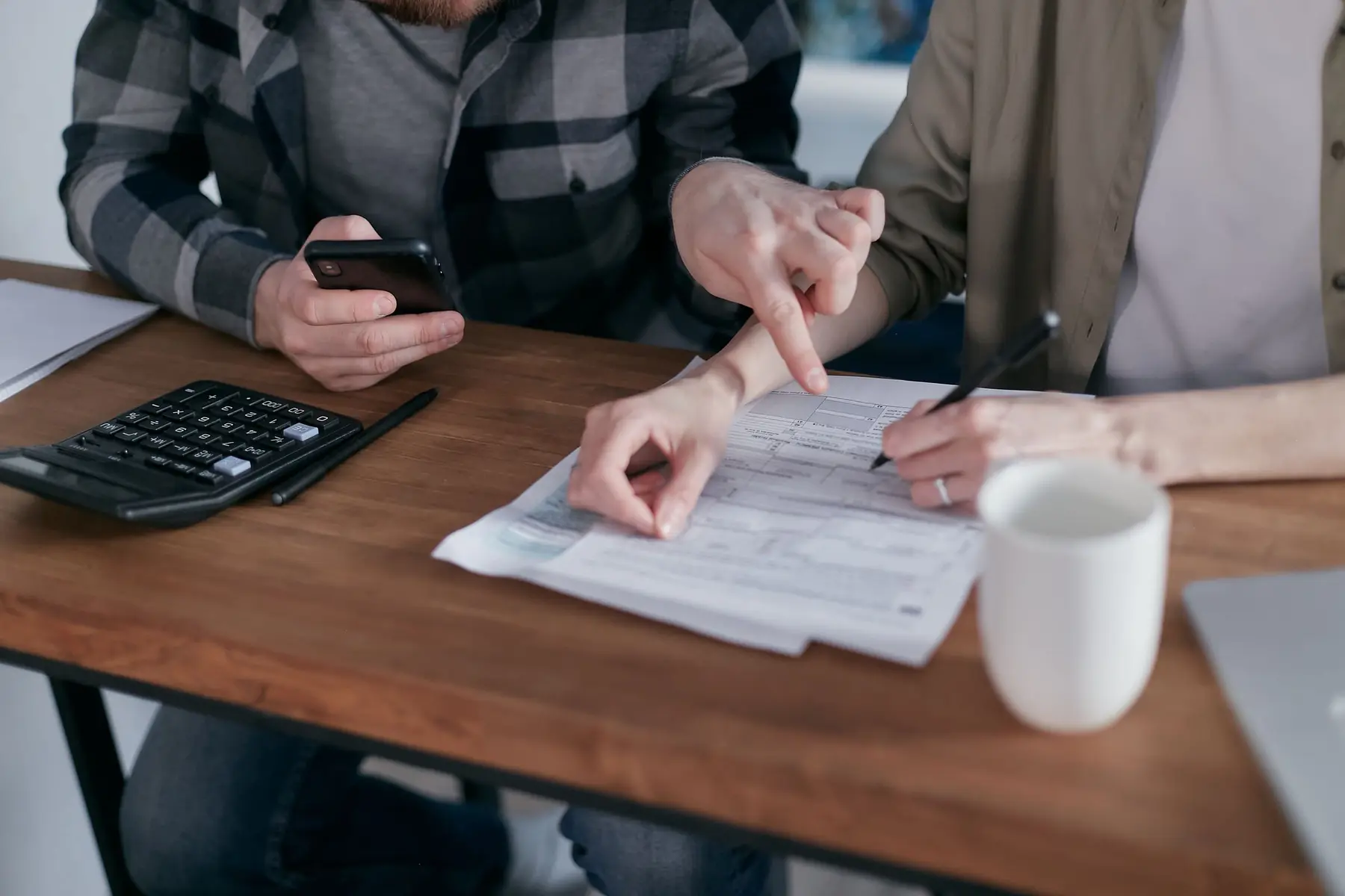 Mãos sobre uma mesa de madeira analisando um contrato de crédito ao lado de calculadora, celular e caneta, representando a conferência do custo real antes de assinar