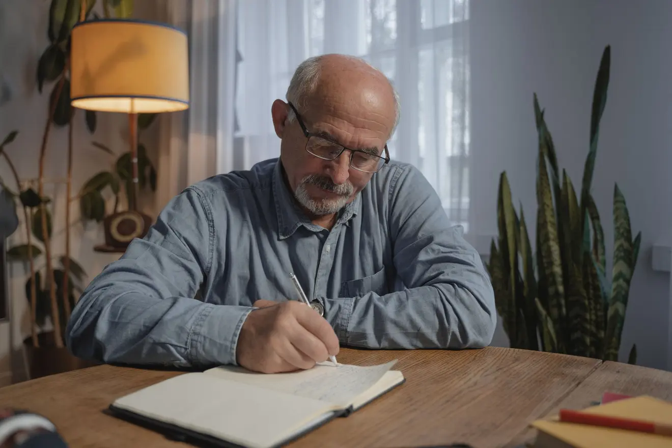 Homem de mais idade escreve em um caderno sobre uma mesa de madeira em casa, iluminado pela luz quente de um abajur