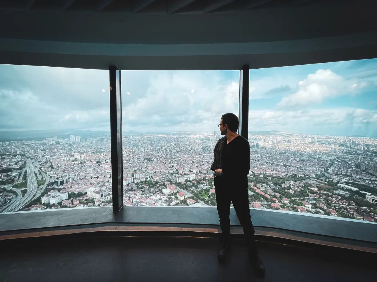 Homem visto de longe diante de uma janela panorâmica curva, observando uma cidade que se estende até o horizonte