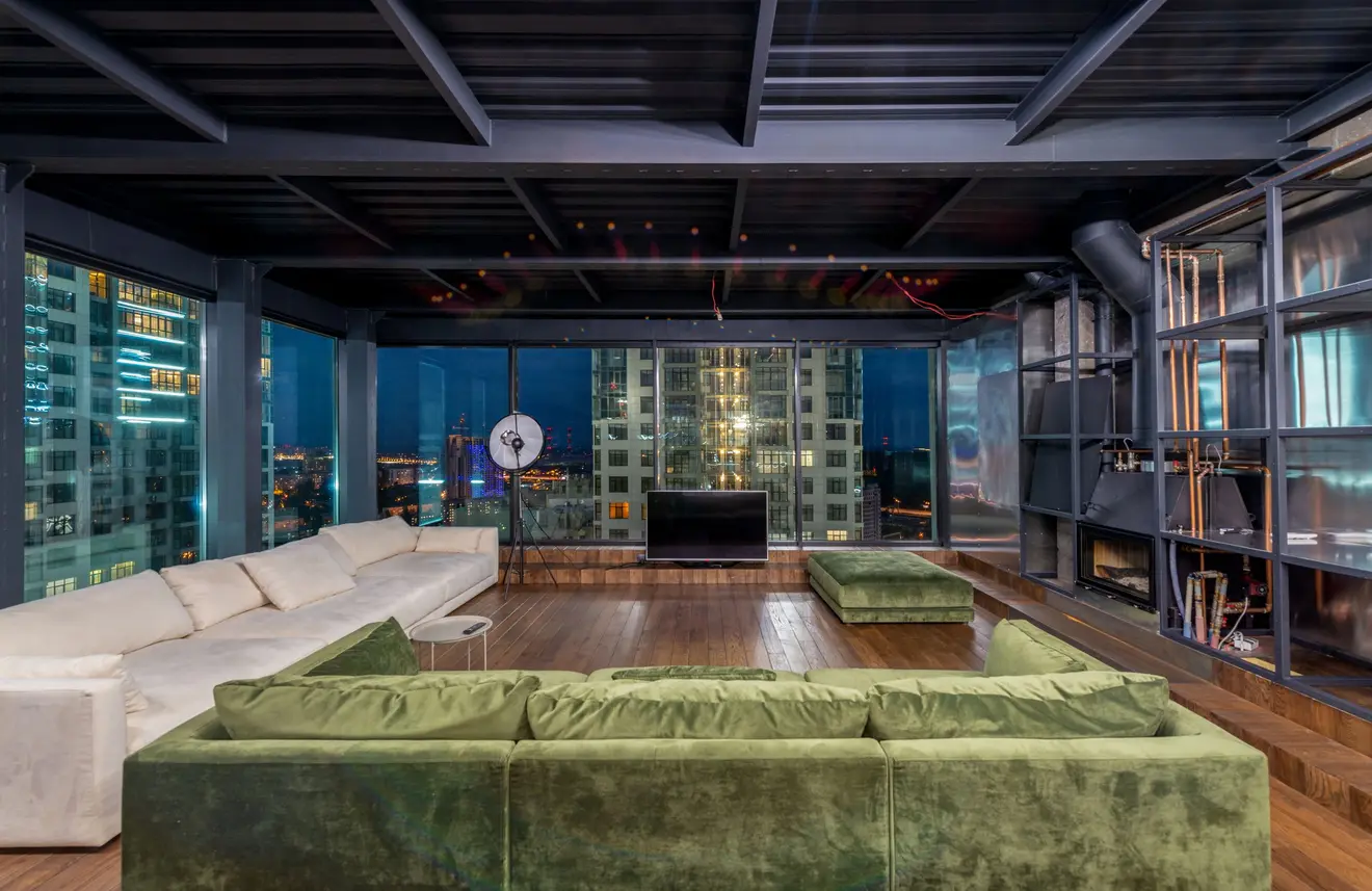 Sala de estar de uma cobertura escura e sofisticada à noite, com sofá de veludo verde e janelas do chão ao teto abrindo para torres iluminadas da cidade