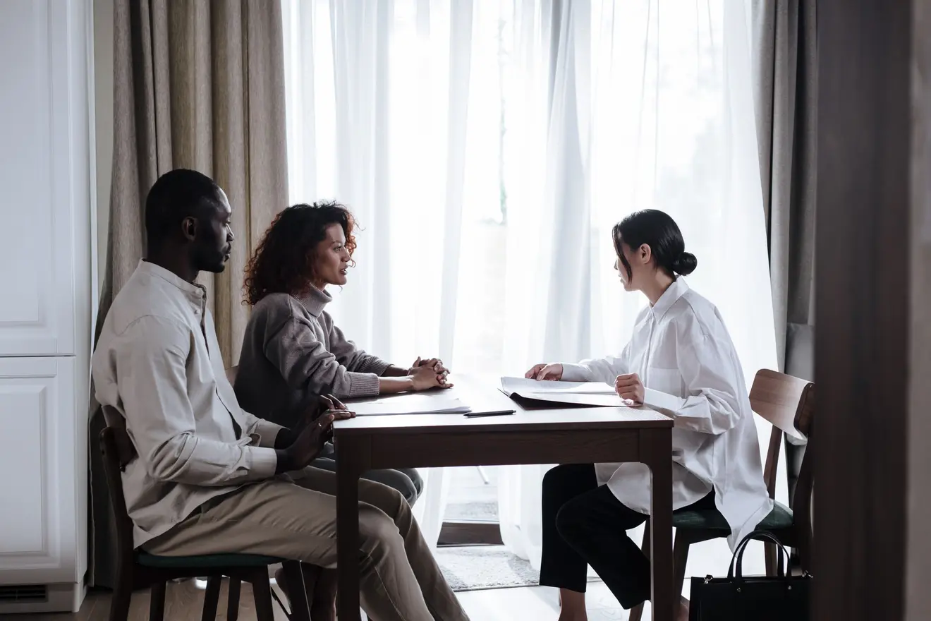 Casal sentado à mesa diante de uma consultora, analisando documentos com seriedade, com luz natural entrando pelas cortinas