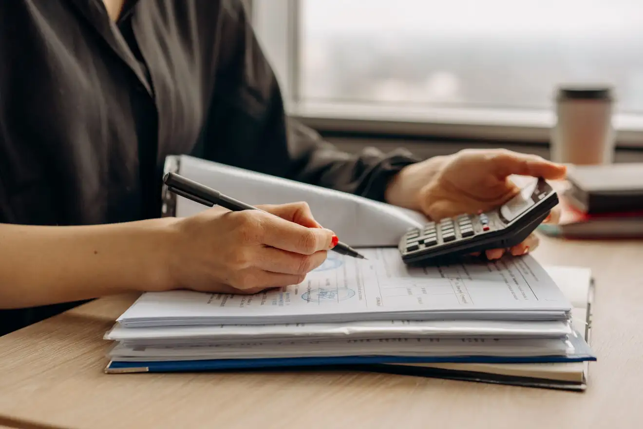 Pessoa em uma mesa com calculadora, caneta e uma pilha de documentos, fazendo as contas do custo real de vender um imóvel