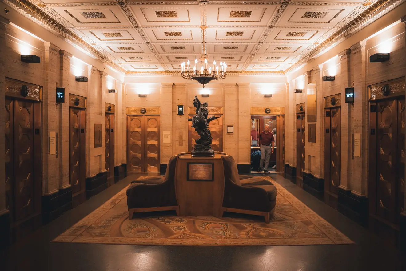 Hall de entrada de um prédio residencial tradicional, com lustre, sofás, painéis de madeira e portas de elevador