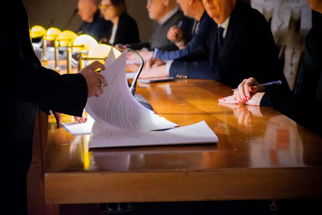 Mesa de reunião em luz baixa: uma mão ergue a página de um contrato diante de uma fileira de executivos de terno do outro lado